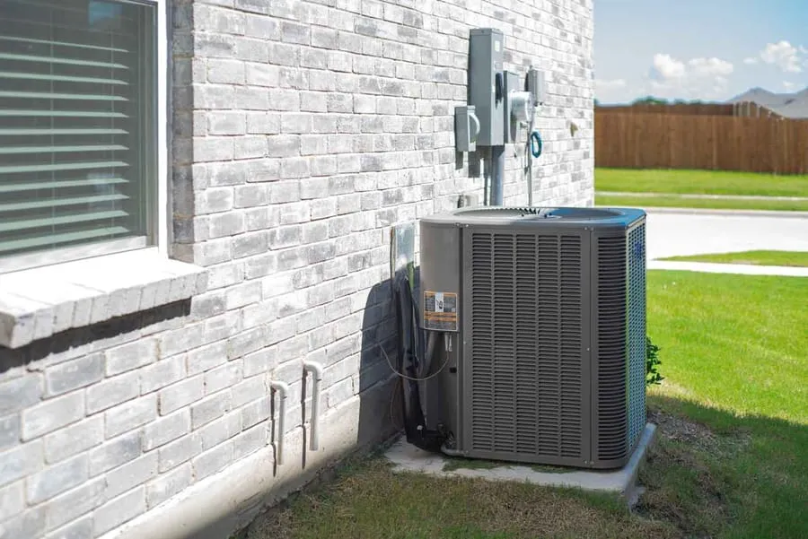 Side windows of new home and central air conditioning condenser unit mounted on concrete slab beside brick house wall